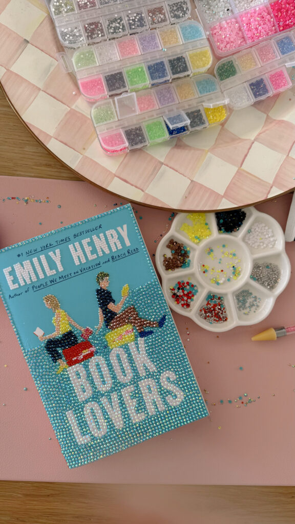 Half bedazzled Book Lovers by Emily Henry book placed on a table alongside different color gems 