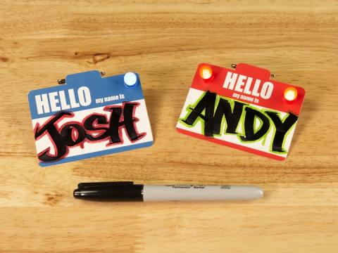 Two LED name tags, one blue with the name Josh and one red with the name Andy, on a wooden surface with a black permanent marker.