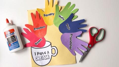 a photo of a rainbow assortment of handprint made from construction paper each with a different inspiration word pasted on to a large sheet on construction paper under neath the handprints is a coffee mug with the text "i have a dream.' The craft is next to a bottle of glue and scissors. 