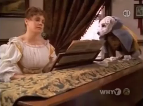 A woman with her hair pulled back wearing a ruffled dress sits at a wooden upright piano, covered in a fabric. A russell terrier dressed in period clothing sits atop the piano.