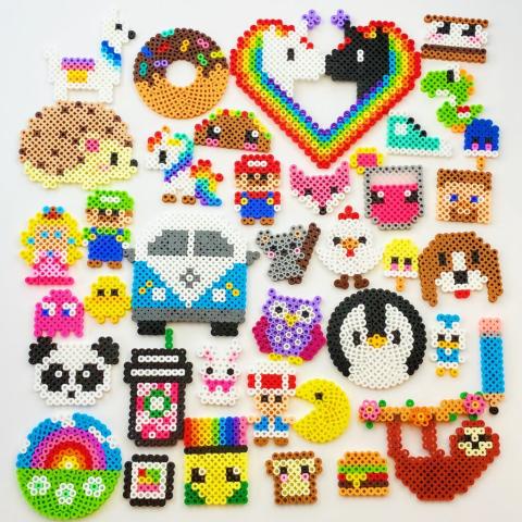 A collection of colorful Perler bead creations arranged on a light background. The designs include pixel-style figures such as animals, food items, rainbows, hearts, and characters. Visible shapes include a panda, penguin, owl, sloth, unicorns, a donut, taco, burger, coffee cup, and a rainbow heart. The pieces are made from small fused plastic beads and feature bright, playful colors and detailed patterns.