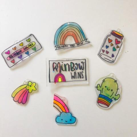 A collection of small, handmade shrink plastic charms displayed on a light surface. The charms feature colorful, hand-drawn designs including rainbows, hearts, a smiling cloud with a rainbow, a cactus in a rainbow pot, a shooting star, and a jar filled with hearts. One charm reads “RAINBOW WINS,” and another includes a rainbow with the phrase “rainbow days ahead.” The designs are bright, playful, and outlined in black.