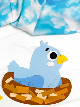 The image depicts a paper craft made of a jubilent bluebird on a brown nest. The nest is composed of varying shades of brown. The craft sits on marble countertop with a unique sky patterned cloth.