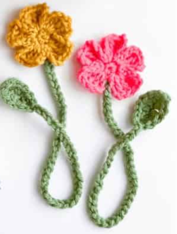 This photo is of two crocheted flowers. Both have green stems with one leaf. The flower on the left has yellow petals, while the flower on the right has pink petals.